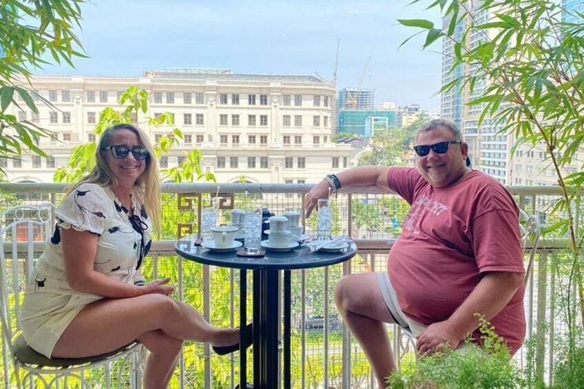 Two people seated at a round table with cups and saucers, outdoors with a cityscape in the background. 5
