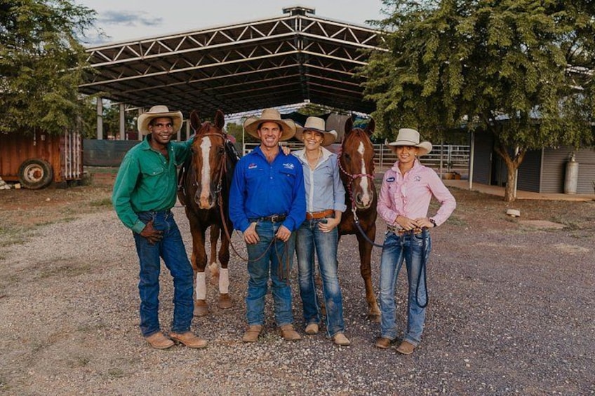 A group of people standing with horses in an outdoor setting with a metal shelter and trees. 2