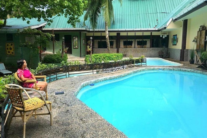 A swimming pool with a person sitting on a chair, surrounded by greenery and a building with a green roof. 4