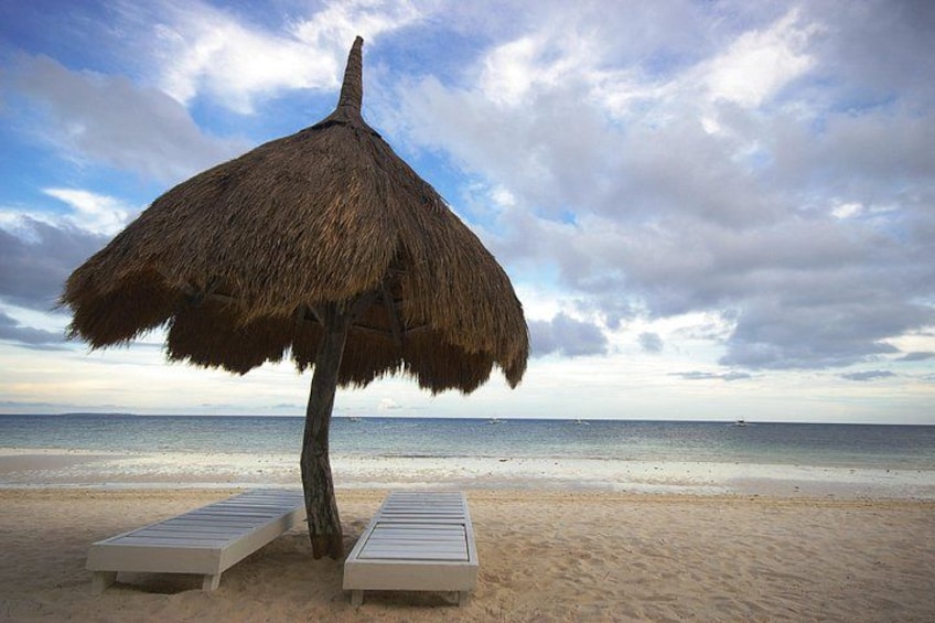 A beach with a thatched umbrella, two lounge chairs, and the ocean in the background. 1