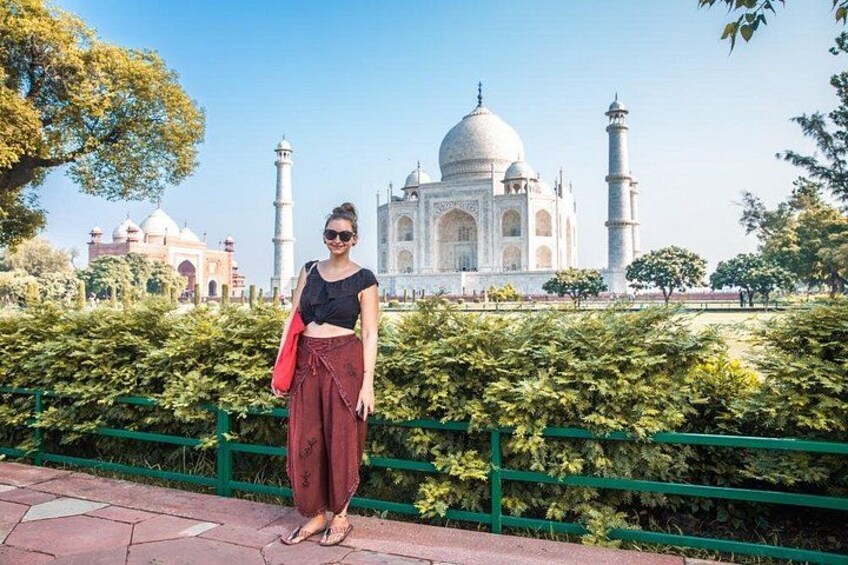 A person standing in front of the Taj Mahal with a green fence and trees in the foreground. 5