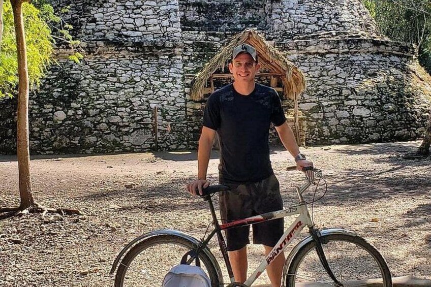 Une personne se tient à côté d’un vélo, avec un mur de pierres et une construction à toit de chaume en arrière-plan. 3