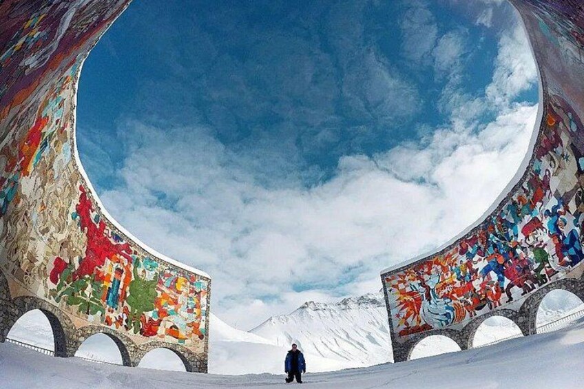 Una persona de pie en un túnel con murales coloridos en las paredes, que conduce a un paisaje montañoso nevado. 3