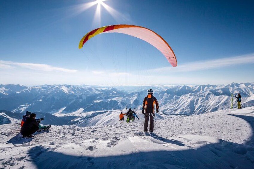 Una persona practicando parapente en la cima nevada de una montaña, con cielo despejado y sol. 4