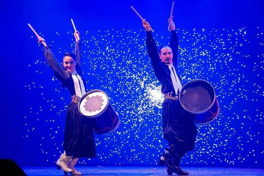 Two performers with drums, one in a black dress and the other in a suit, on a stage with a blue background and sparkling lights. 3