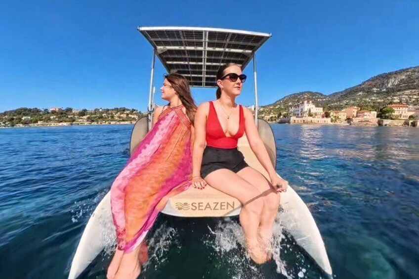 Two people on a boat with a canopy, water, and a coastal landscape with buildings. 4
