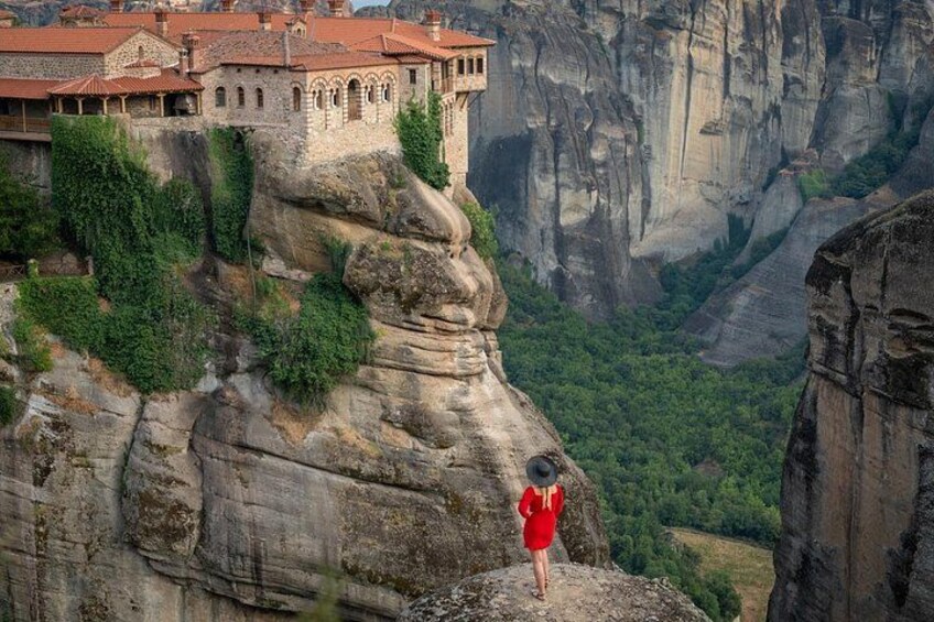 A person in a red dress standing on a rock ledge overlooking a valley with a monastery on a cliff. 4