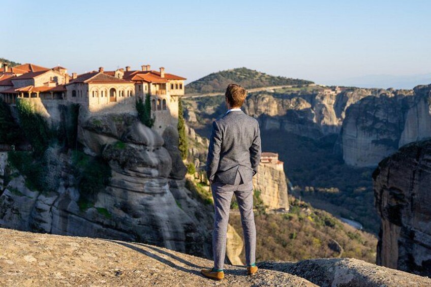 A man in a suit standing on a rocky outcrop overlooking a valley with a building on a cliff. 2
