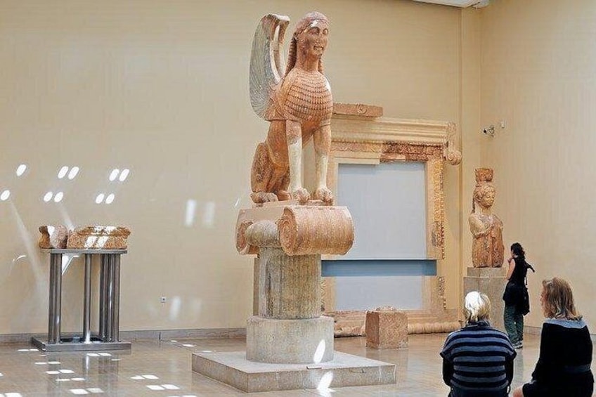 A museum interior with a large statue of a winged figure on a pedestal, a display case with artifacts, and a wall with a relief sculpture. 3