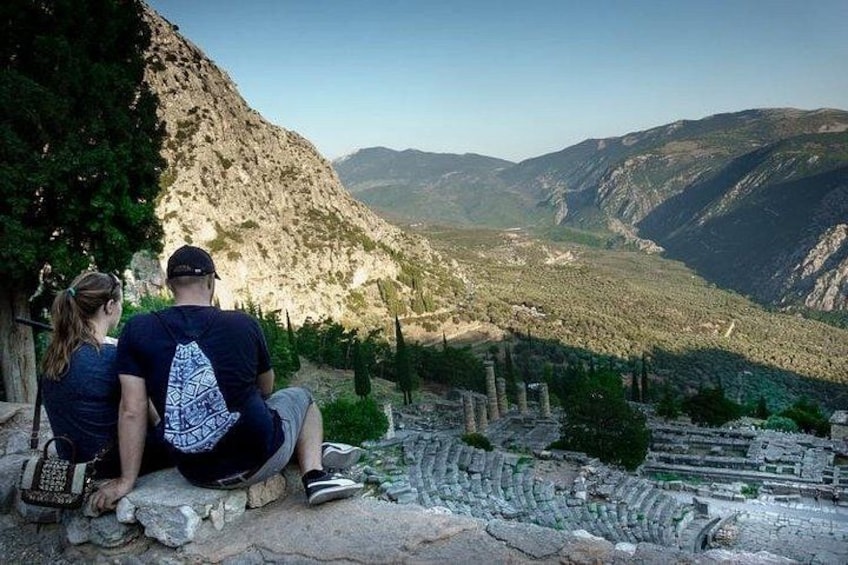 A couple sitting on a stone ledge overlooking a valley with ruins and mountains in the background. 2