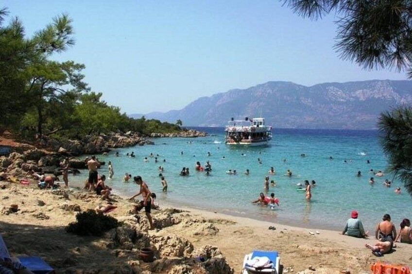 Una playa con gente nadando y tomando el sol, un bote en el agua y montañas al fondo. 3