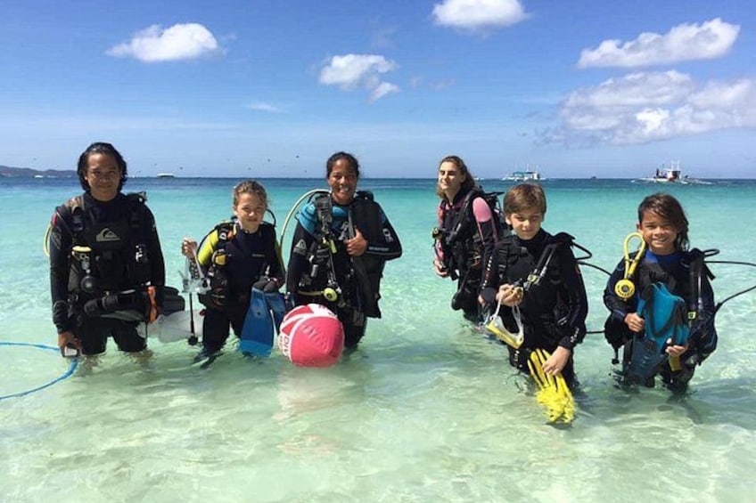 Un groupe de personnes, équipées de matériel de plongée sous-marine, se tient dans une eau peu profonde, avec un ciel dégagé et une mer calme en arrière-plan. 1