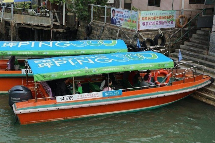 Een boot met een blauw dakzeil en de tekst 'PING' erop, afgemeerd aan een steiger met een trap naar een gebouw. 3