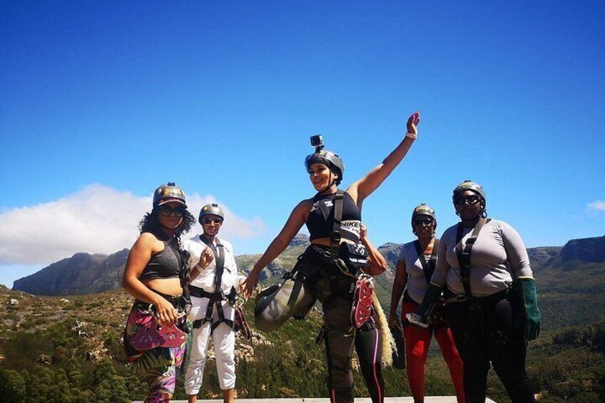 A group of people with backpacks and helmets, standing on a mountain with a clear sky and a mountainous landscape in the background. 4