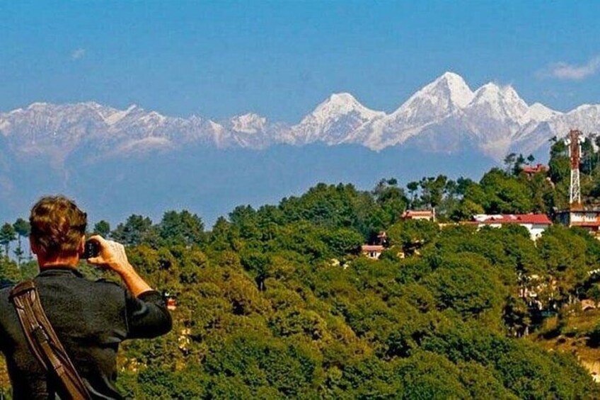 A person taking a photograph of snow-capped mountains with a forested hill in the foreground. 3