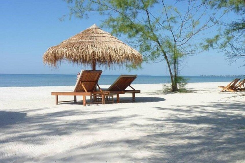 A beach with two lounge chairs under a thatched umbrella, a sandy shore, and a clear blue sky. 1