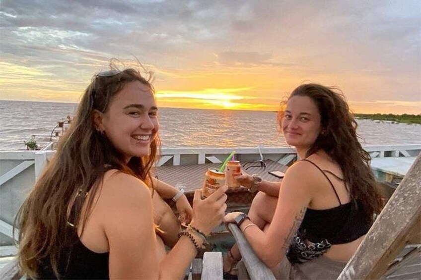 Twee personen zitten op een steiger, drankjes vasthoudend, met een zonsondergang op de achtergrond. 1