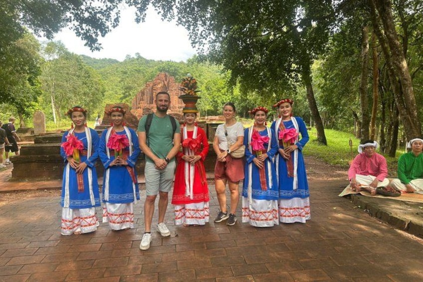 Eine Gruppe von Menschen in traditioneller Kleidung steht in einem Park, im Hintergrund ein Tempel. 1
