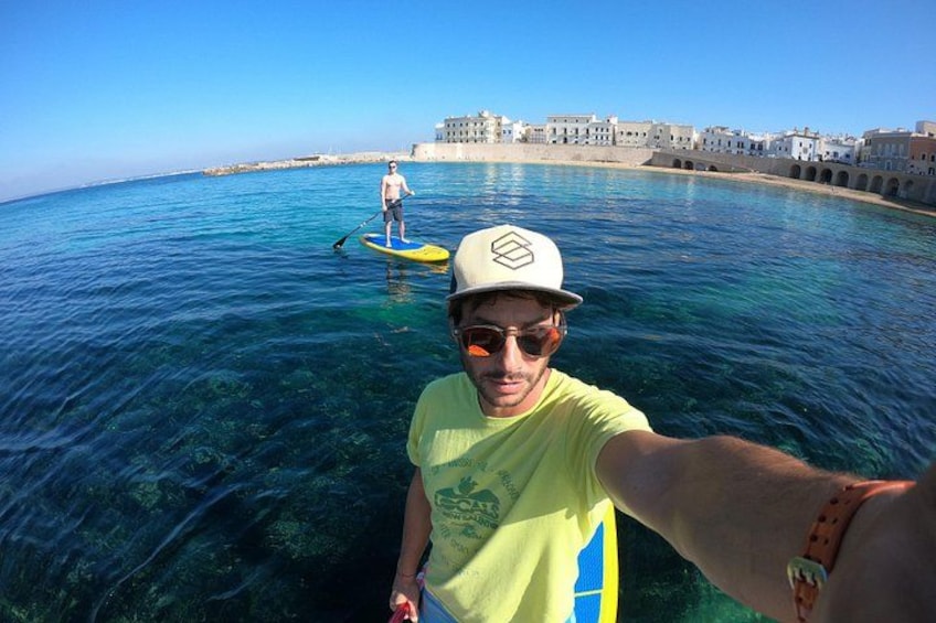 A person paddleboarding on clear blue water with a coastal town in the background. 1