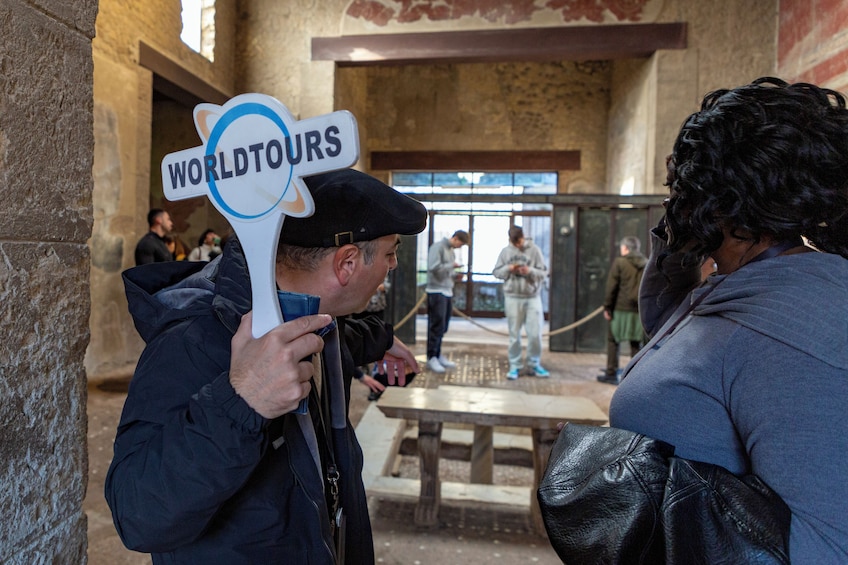 Un hombre sostiene un letrero que dice 'WORLDTOURS' en un edificio histórico con paredes de piedra y una mesa de madera. 3