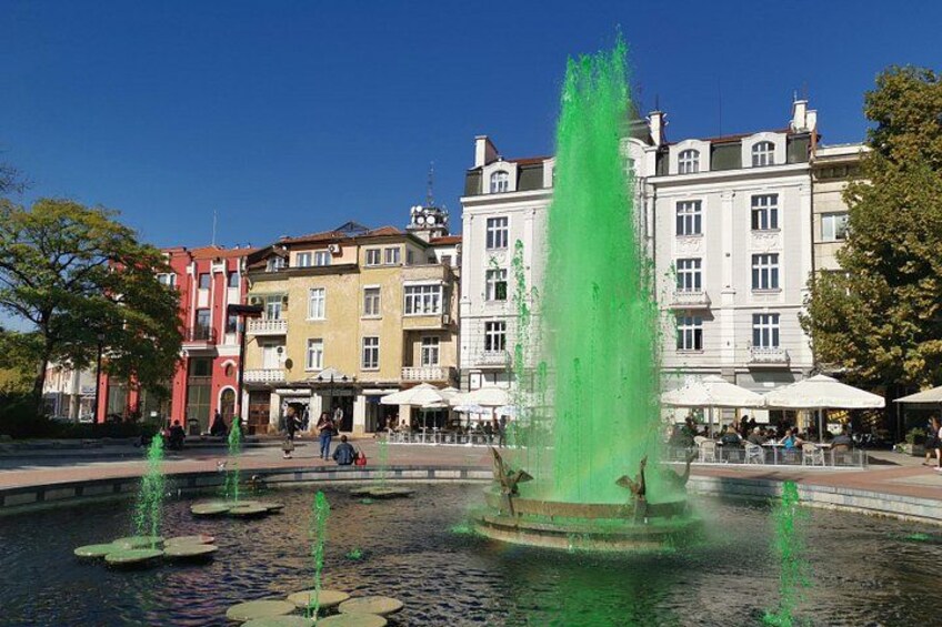 A fountain with water jets in a square, surrounded by buildings and trees. 3