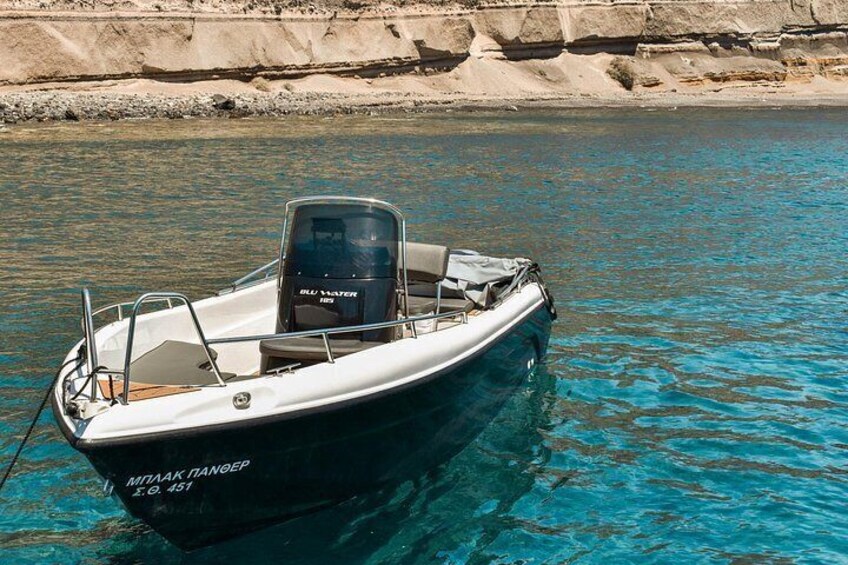 A small motorboat named 'Mitaak Nandeep' with a motor and seating, docked at a calm body of water with a rocky shoreline in the background. 1