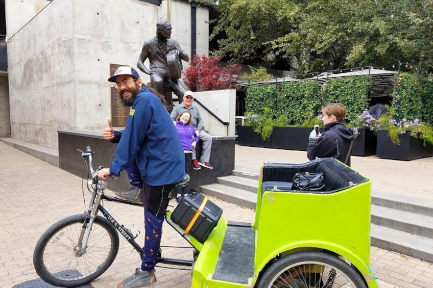 A person on a bicycle with a bright green cargo bike, a statue, and a building with greenery in the background. 2