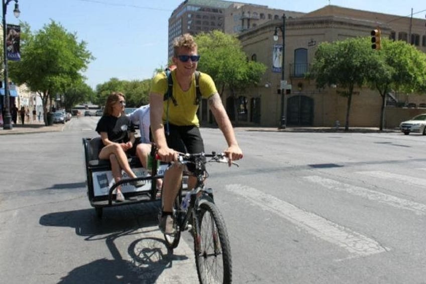 Una persona pedaleando en bicicleta con un remolque, otra persona sentada en el remolque, y un edificio al fondo. 3