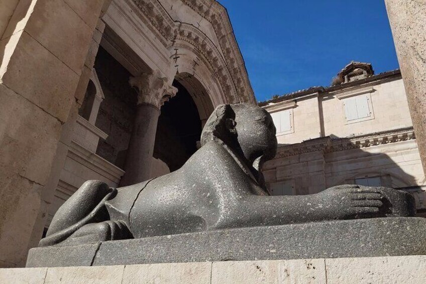 Una estatua de esfinge de piedra en un entorno exterior, con arquitectura clásica al fondo. 1