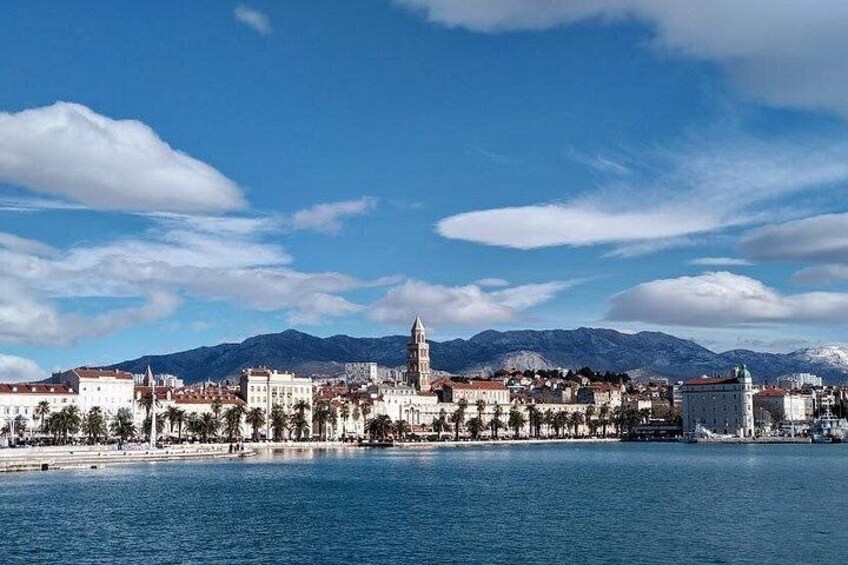 Un paisaje urbano costero con una torre destacada, mar tranquilo y montañas al fondo. 4