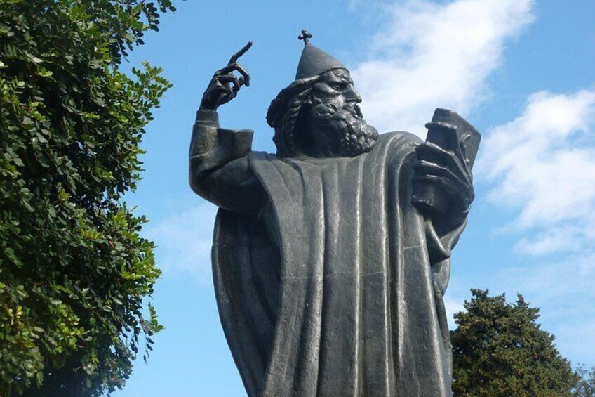 Una estatua de una figura vestida con túnica, con un dedo señalando, sosteniendo un libro y usando un sombrero. 5