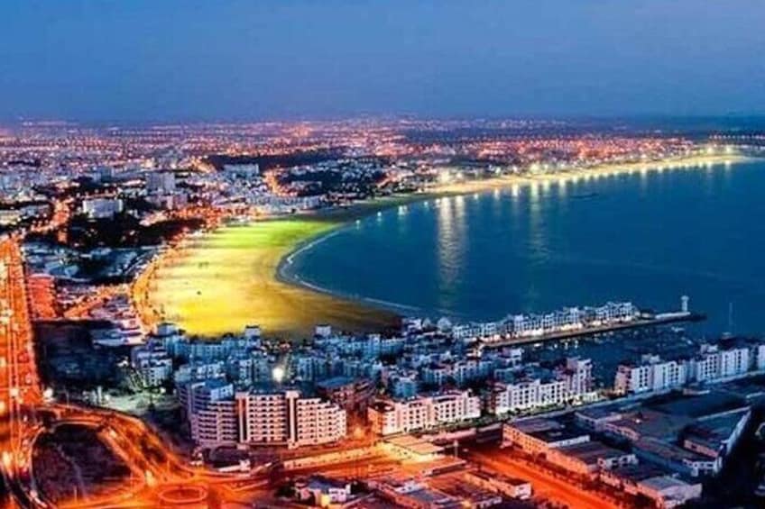 Une vue d’une ville côtière au crépuscule, avec une plage, des bâtiments illuminés et un port de plaisance. 5