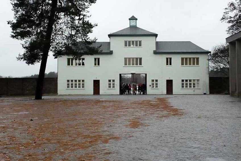 A white building with a grey roof, a large open entrance, and a paved courtyard. 1