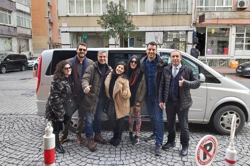 A group of people posing in front of a silver van on a cobblestone street with buildings in the background. 4