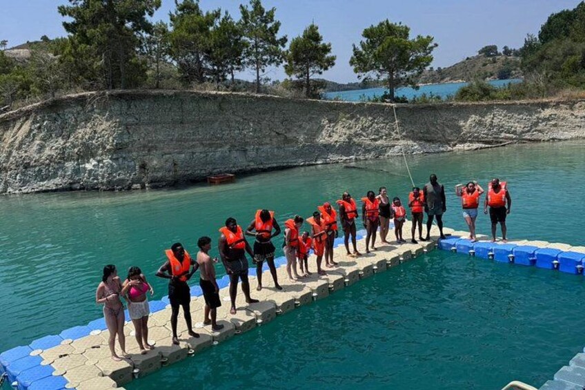 A group of people in orange life jackets standing on a dock by a clear blue pool. 3