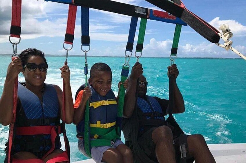 Tres personas en un bote con chalecos salvavidas, un mar azul cristalino y una vela blanca al fondo. 2