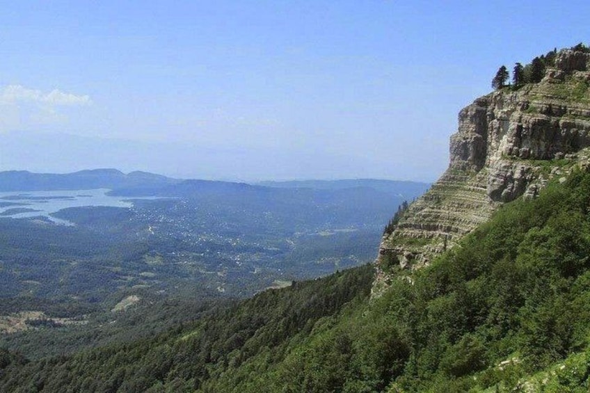 A mountainous landscape with a steep cliff, dense forest, and a distant body of water. 2