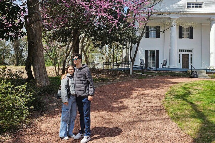 A couple standing in front of a white building with black shutters, surrounded by trees with pink blossoms. 2