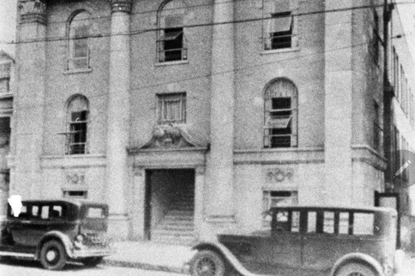 A building with arched windows and a central entrance, a vintage car parked in front, and a truck to the right. 5