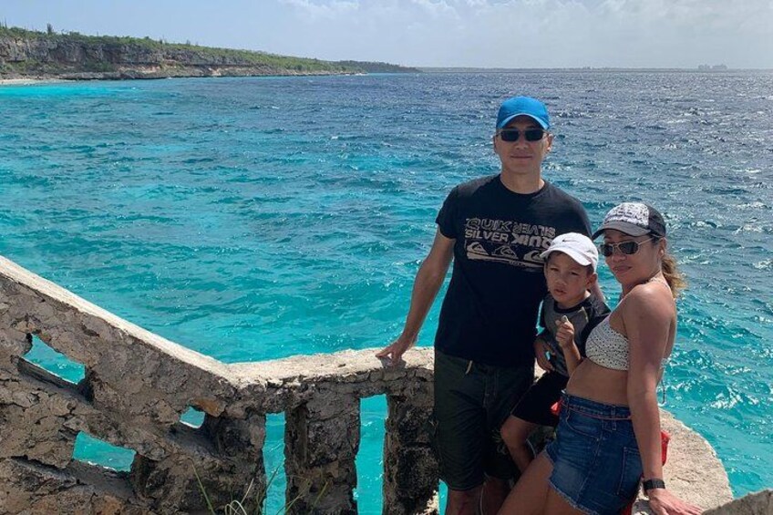 A family of three by a turquoise sea with a rocky shoreline. 2