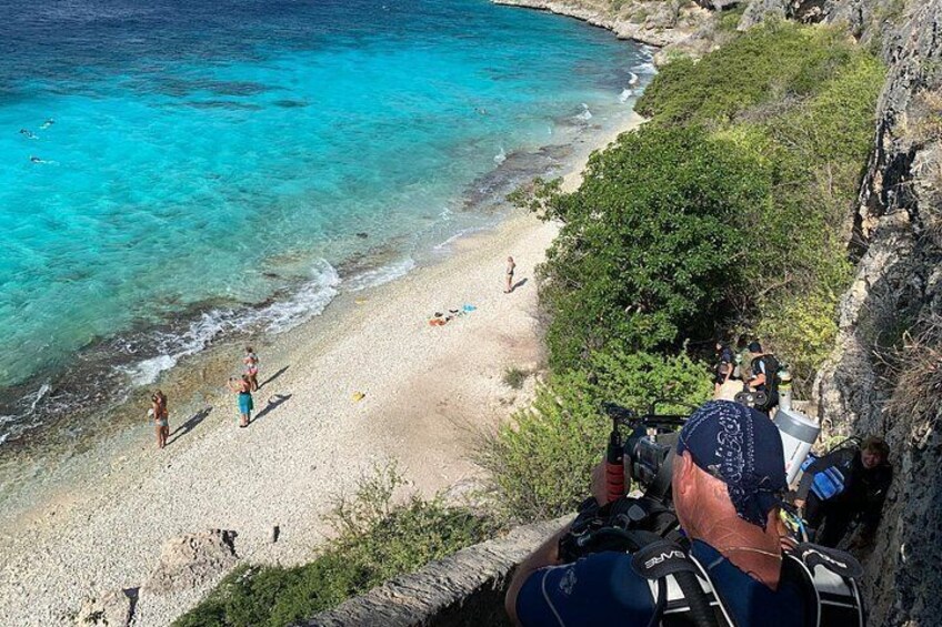 Una spiaggia con acqua turchese cristallina, persone che camminano sulla riva e una scogliera rocciosa in primo piano. 3