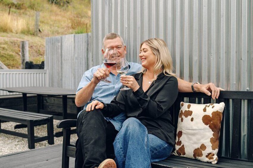 Two people sitting on a bench outdoors with a wooden fence and a cow-patterned pillow in the background. 3