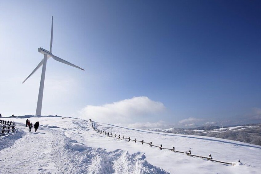 A large wind turbine on a snowy hill with a fence and people walking. 3