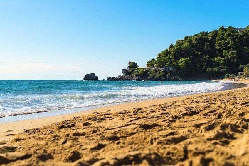 A sandy beach with clear blue water, rocky outcrops, and dense green foliage in the background. 2