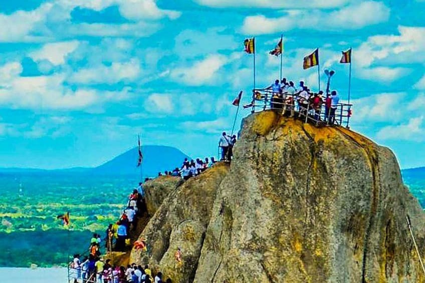 Ein felsiges Vorsprungs mit Personen darauf, Flaggen und eine gebirgige Landschaft im Hintergrund. 3