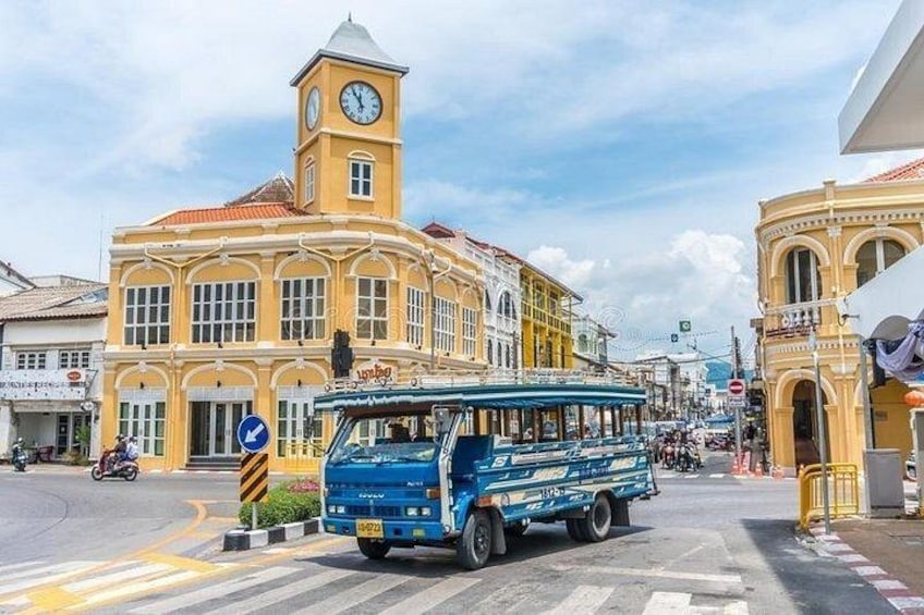 A blue bus at a roundabout with a clock tower and yellow buildings. 3