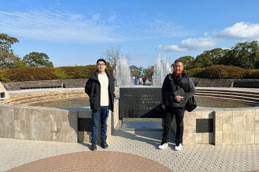 Two people standing in front of a fountain with a stone wall and a plaque. 4