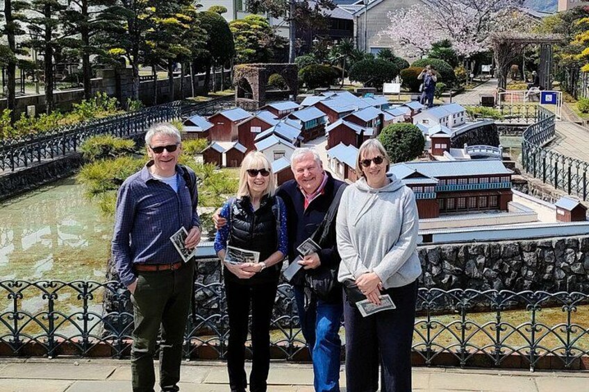 A group of people standing by a model village with a water body in the background. 5