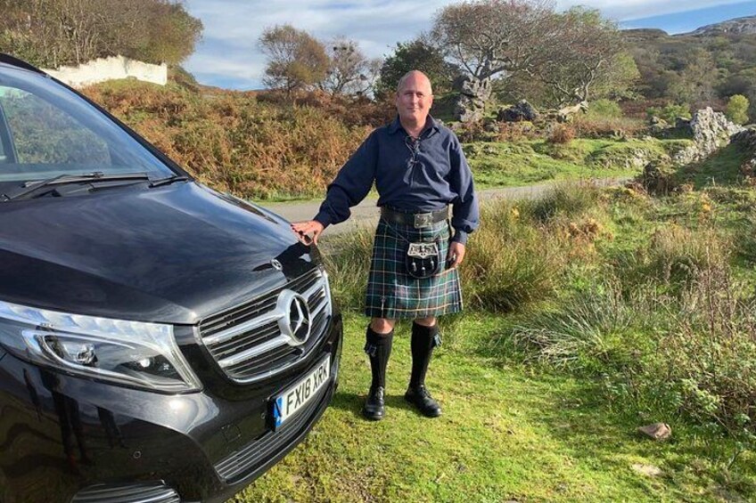 A person in a kilt standing next to a black Mercedes-Benz car. 1