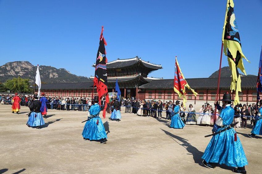 A traditional dance performance with participants in blue dresses, flags, and a traditional building in the background. 1